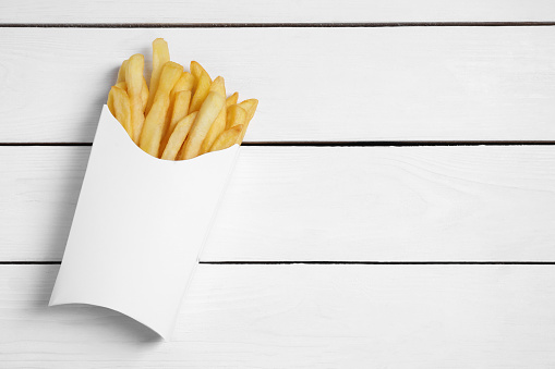 Delicious french fries in paper box on white wooden table, top view. Space for text