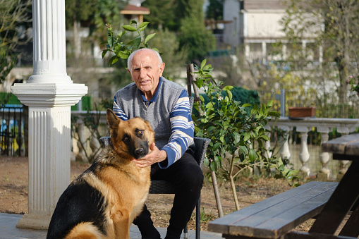 Portrait of a smiling old man sitting in his garden with his dog. Old man relaxing outside with his dog and both looking at the camera. Photo of a happy old man having a good time with his dog in his garden.