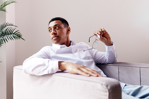 serious pensive african american man in glasses and shirt sits in an easy chair and looks to the side out the window, guy in business clothes holds glasses and rests in comfortable chair