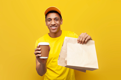 young african american delivery man in yellow uniform holds order in package on yellow background, service worker gives order
