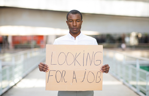 Unemployed black man wearing shirt holding placard with looking for a job text placard, upset unhappy african american male lost his work, suffering unemployment crisis, posing outdoors, copy space