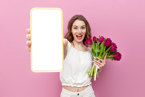 young woman in white festive clothes holds bouquet of pink tulips and shows blank smartphone screen on isolated background, girl with flowers advertises phone, concept of spring and women's holiday