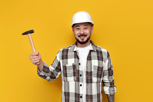 asian builder in hard hat and shirt holds hammer on yellow isolated background, korean worker constructor with tool in goggles