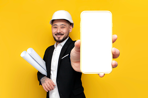 asian foreman in black suit with papers and projects shows blank smartphone screen on yellow isolated background, korean civil engineer in hard hat and glasses advertises phone mockup