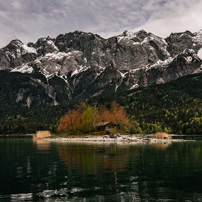 A small wooden house is situated in the serene waters of a pristine lake surrounded by majestic snow-capped mountain peaks
