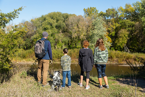 Rear view of a father, his three children, and their pet dog, an Australian Shepherd out in nature on an autumn day in Minnesota, USA. They are standing on the bank of the Crow River at Crow-Hassan Park Reserve.