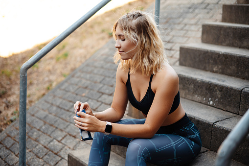 Young fitness woman in sportswear opens a fitness bar while sitting on stairs outdoors. Blonde girl resting after workout outdoor. Healthy and sport concept