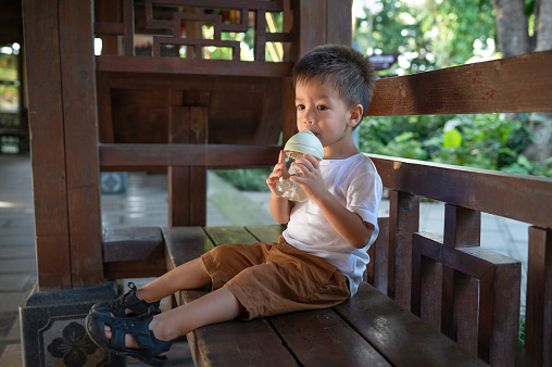 Charming two and a half year old boy enjoys juice in one of China's beautiful parks. During a break between playing and having fun outdoors, an energetic boy resting on a wooden bench with a healthy juice