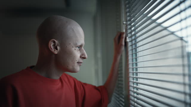 SLO MO Side View of Upset Young Bald Man with Cancer Looking Through Window Blinds at Home