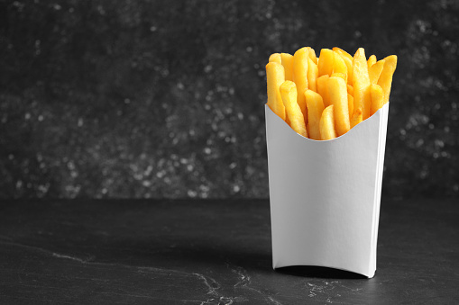Delicious french fries in paper box on black table, space for text