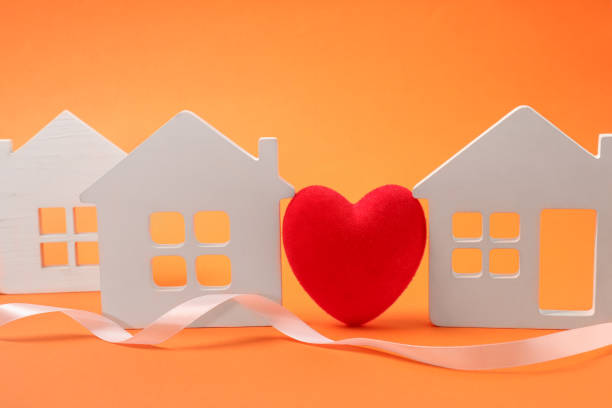 istock Long-distance relationship concept. Decorative heart between white house models and ribbon on orange background 1923286749