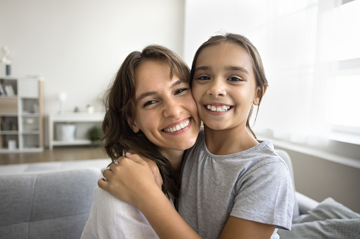 Cheerful beautiful mom and happy tween daughter kid hugging with love, faces touch, looking at camera, smiling, showing white teeth, posing for home close up portrait, talking on video call