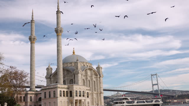 Handheld Shot of Flock of Birds Flying over Ortakoy Mosque with Bosphorus Bridge in Background against Cloudy Sky at Istanbul,Turkey