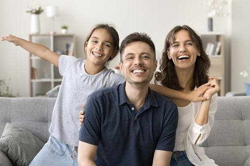 Cheerful happy parents and kid with open flying arms having fun on home couch, looking at camera with toothy smiles, enjoying family leisure, together, laughing. Video call screen, head shot portrait