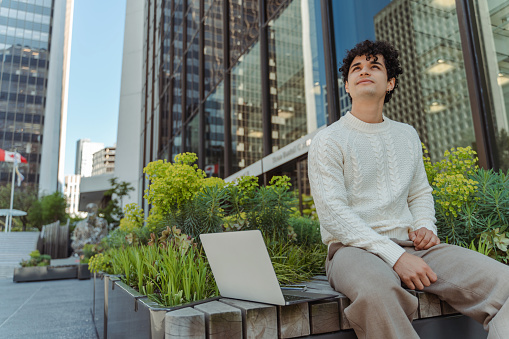 Hispanic smiling businessman looking away and resting after work outdoors. Portrait of handsome successful latin freelancer check email sitting at workplace. Technology concept