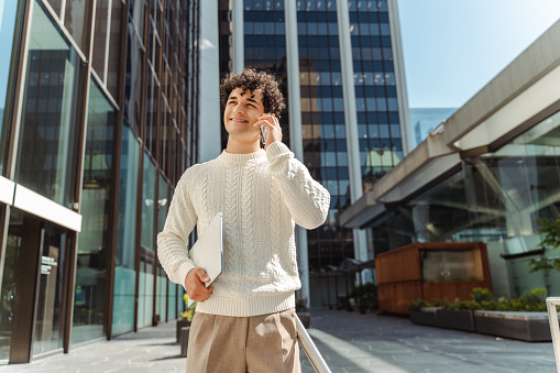 Portrait of handsome smiling Latin man talking on mobile phone, answering call holding laptop looking away walking on urban street, copy space. Successful business, technology concept