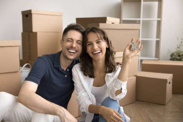 Cheerful excited young couple happy to move into new house Cheerful excited young couple happy to move into new house, showing keys, hugging at stacked cardboard boxes, looking at camera, smiling, laughing, posing for portrait happy family with key of their new house stock pictures, royalty-free photos & images