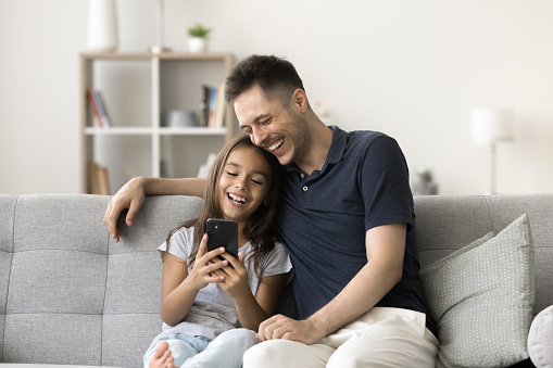 Happy joyful dad and little daughter child using online communication on smartphone, using digital device together, resting on home couch, enjoying leisure, relationships, fatherhood