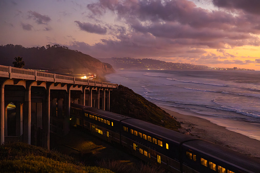 Close-up of a train passing on the tracks under an overpass near Torrey Pines State Beach at San Diego. It is dusk, and the illuminated windows of the train give the picture a special charm