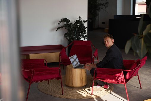 Man working laptop in cozy and comfortable corner of big working zone of coworking space, sitting in red armchair and looking at camera, hourly payment and free wi-fi