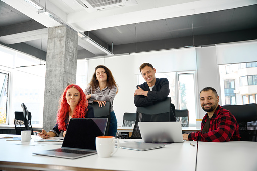 Positive and friendly team of male and female managers staying on workplaces with laptops and looking at camera with smile, posing to corporate photo, high-qualified professionals