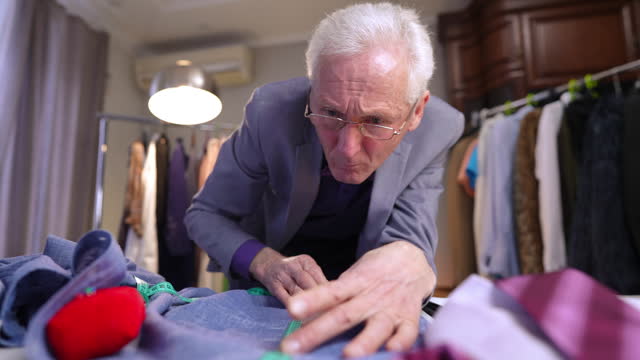 Concentrated, the man marks a line with white chalk on the fabric of his blue shirt. An elderly fashion designer works in the workshop.