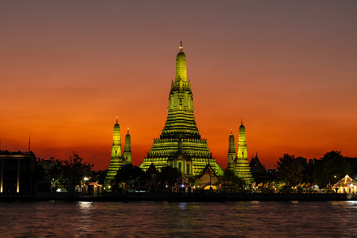 Wat Arun (Temple of the dawn) in Bangkok at dusk. Temple and prangs bathed in yellow light. Deepening orange sky in background. Chao Phraya river in foreground.