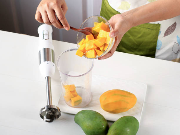 Female Chef hand Pour Diced Mango to the Juicer Bowl, Process Making Mango Smoothie or Mango Lassi in the Kitchen stock photo
