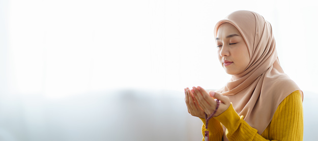 Asian beautiful Muslim woman in in Hijab dress sitting on the floor and praying for Allah in her house. Muslim lifestyles concept.