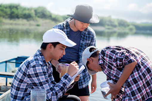 Asian boys in plaid shirt hold transparent plastic bottles and  cup which had water from river in front of them inside, they are studying water, environment, river, insects, particles and eco effect.