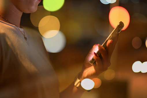 Closeup of female hands holding mobile phone browsing internet with blurred lights at night.
