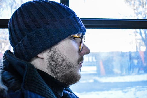 portrait of a young man, he is sitting by the window, he has a knitted cap, he wears glasses, he has a beard, it can be seen from the profile