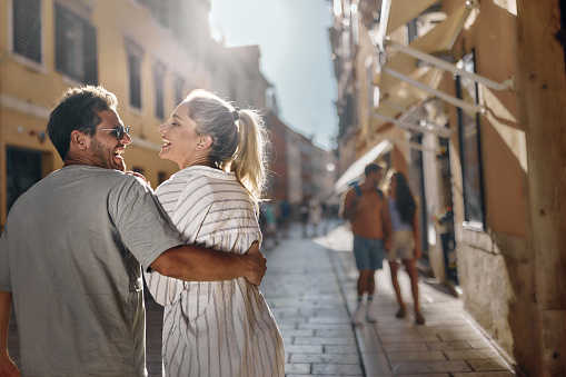 Back view of cheerful couple talking while walking embraced during spring day on the city street.