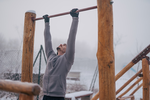Strong young man doing pull-ups on outdoor bar while training at workout area in winter