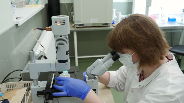 Woman looks through microscope in laboratory. Stock footage. Masked woman looks at biochemical substance through microscope. Analysis of substance under microscope in laboratory.
