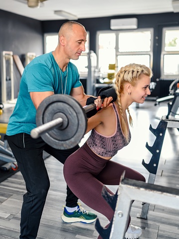 Young woman doing barbell squats with the assistance of a trainer.