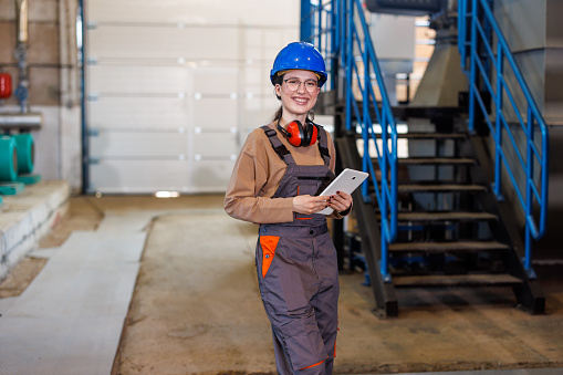 This photo captures a woman performing checks on a digital tablet while standing in a heating plant, with numerous machines and tools in the background