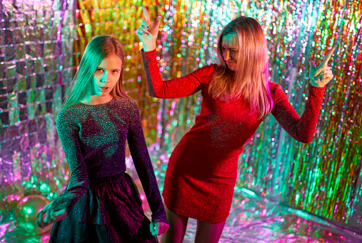Beautiful fair-haired girl dressed in cocktail dress and cute slender middle-aged woman in a tight red dress are daughter and her mom. They are dancing a modern dance together. Studio shooting on a disco party on a glittering background