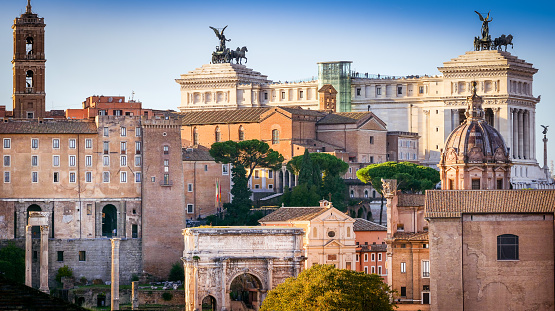 An idyllic cityscape of the Roman Forum seen from the gardens of the Palatine Hill, in the heart of the Eternal City. On the horizon the Altare della Patria National Monument and the bell tower of the Campidoglio or Roman Capitol, on the Capitoline Hill. Bottom center, the triumphal arch of Septimius Severus, along the Sacred Way, and the facade of the church of San Giuseppe and the Mamertine Prison, where the Holy Apostles Peter and Paul were locked up. The Palatine Hill, one of the seven ancient hills of Rome, it is the place where important imperial palaces stood, such as the Domus Flavia and the Domus Augustana. In 1980 the historic center of Rome was declared a World Heritage Site by Unesco. Image in original 16:9 ratio and high definition quality.