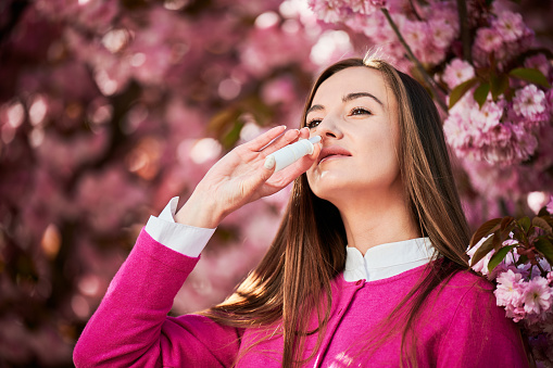 Woman allergic using medical nasal drops, suffering from seasonal allergy at spring in blossoming garden. Close up of woman treating runny nose near blooming tree outdoors. Spring allergy concept.