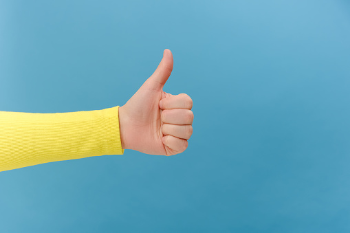 Close up of female hand showing like or thumb up, approval sign, satisfied with service, good feedback concept, isolated on plain blue color background wall in studio with copy space for advertisement