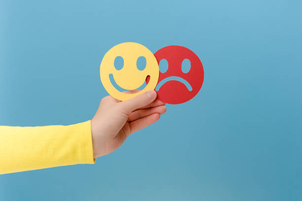 Close up of female hand holding happy and angry face paper, isolated on blue studio background. Feedback rating, emotional intelligence, control, mental health assessment, bipolar disorder concept stock photo