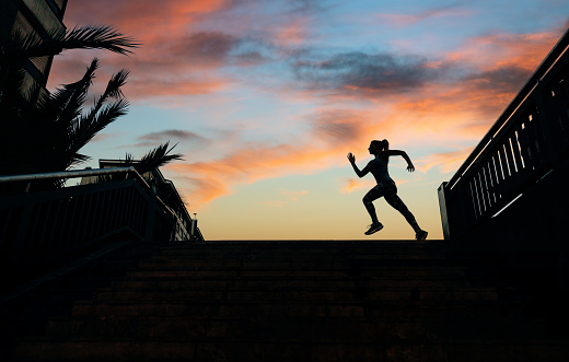 Unrecognizable young female runner silhouette running over an urban background with palm trees and cloudy sunset sky