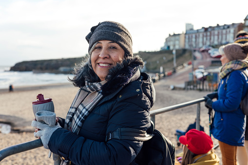 A portrait of a mature female standing outdoors on a beach while enjoying a day out in Tynemouth, North East England. She is looking at the camera smiling while holding an insulated drinks container.