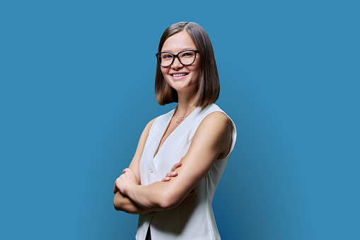 Young confident trendy smiling woman in eyeglasses with crossed arms looking at camera on blue studio background. Beauty, business, work entrepreneurship business expert owner agent concept