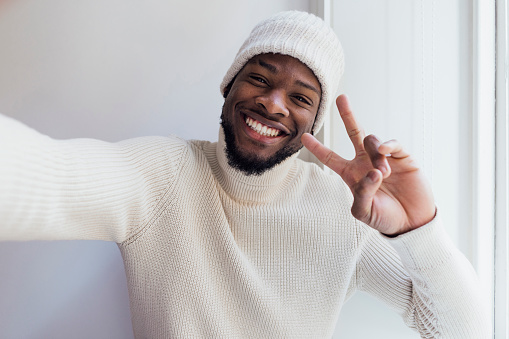 A selfie shot of a young man wearing a cream turtleneck sweater and cream knitted hat, standing next to a window in a building in Newcastle upon Tyne. He is looking at the camera and smiling while posing with the peace sign.