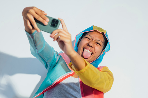 A studio shot of a young woman wearing a multi-coloured raincoat, standing in front of a white background in a studio in Newcastle upon Tyne. She is taking a selfie on a mobile phone while looking at the phone and sticking her tongue out.