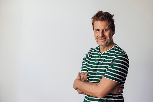 A studio shot portrait of a mid adult man wearing a striped t-shirt, standing in front of a white background with his arms crossed while looking at the camera and smiling. The studio is in Newcastle upon Tyne.