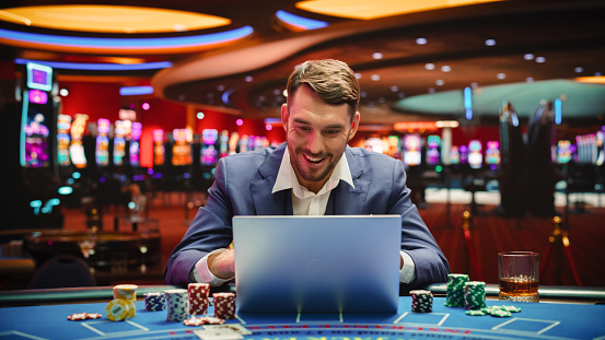 Portrait of a Happy Young Man in a Suit Using a Laptop Computer in Casino. Lucky Businessman Celebrating and Cheering Blackjack Jackpot Win in Online Game on Betting Online Casino Website.