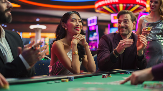Evening in a Stylish Modern Casino: Diverse Group of People Playingh in a Poker Game. Confident Asian Woman Feeling Empowered as She Wins and Receives Admiration and Claps from Players.